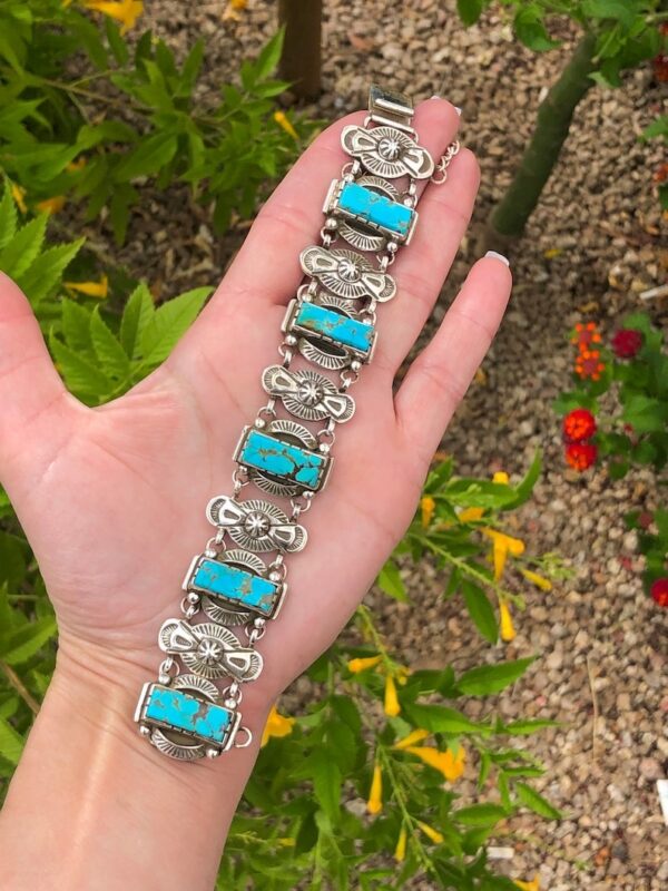 A hand holding a turquoise bracelet in front of some flowers.