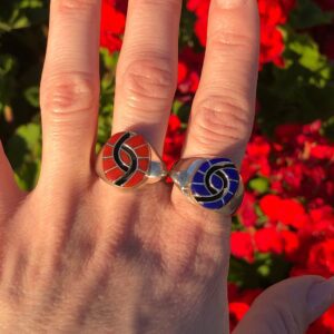 A woman's hand with two AMY QUANDELACY Coral and Lapis Sterling Silver Zuni Rings in front of flowers.