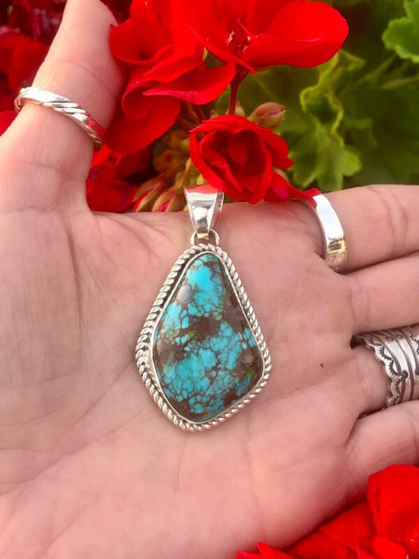 A hand holding a turquoise pendant with red flowers in the background.