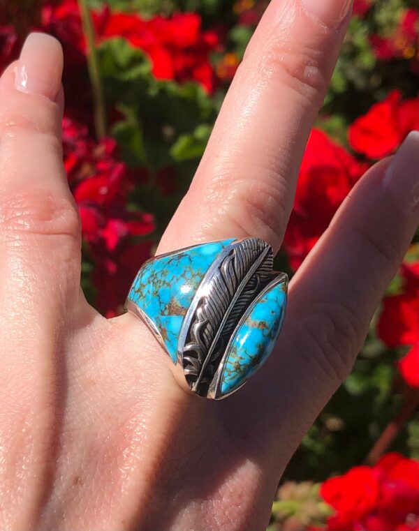 A hand wearing a silver ring with a turquoise feather.