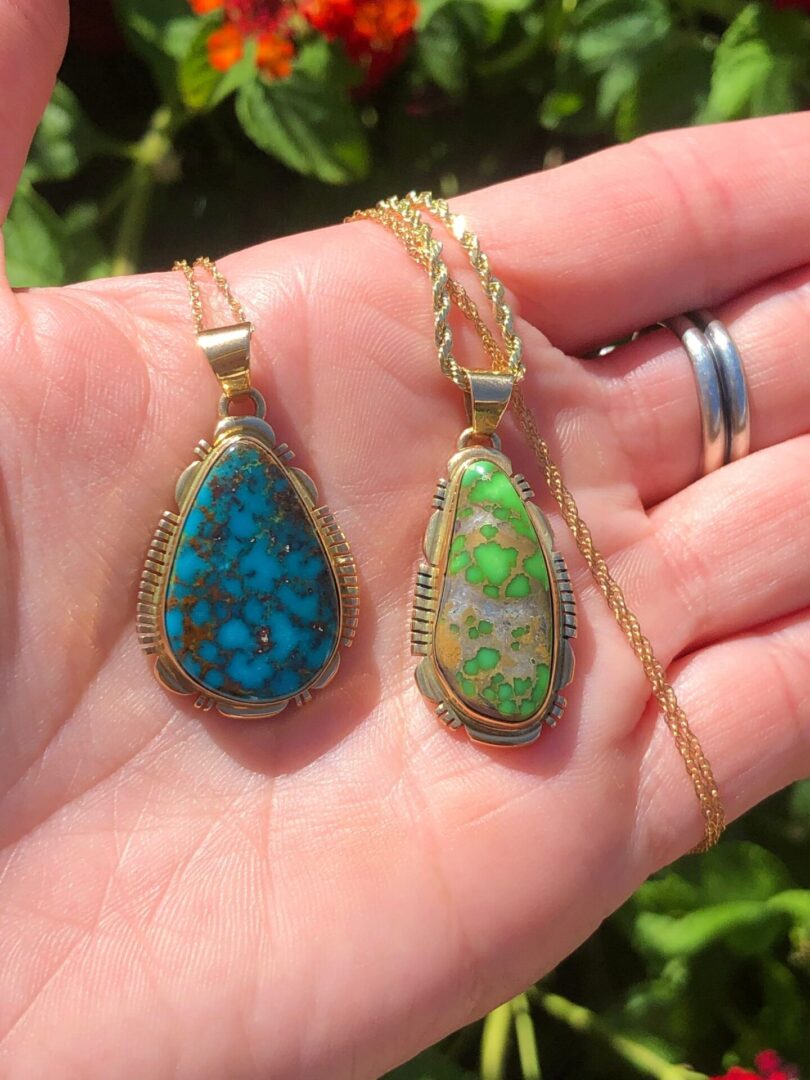 A hand holding the 14KT GOLD AAA Grade Kingman Turquoise + Lime Green Carico Lake Turquoise Navajo Handmade Solid Gold Pendents/Gold Native American jewelry with different colored stones.