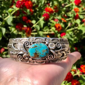 A hand holding a silver cuff with a turquoise stone.