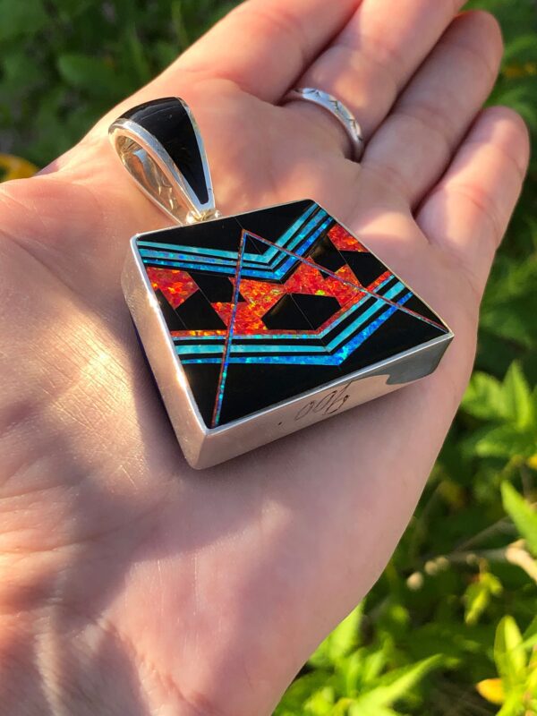 A hand holding a square pendant with red, orange, blue, and black inlay.