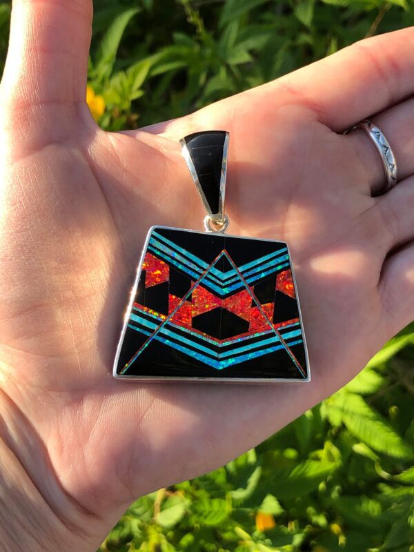 A hand holding a silver pendant with red, orange, blue and black gemstones.