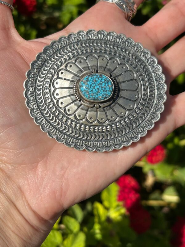 A hand holding a silver belt buckle with turquoise stones.