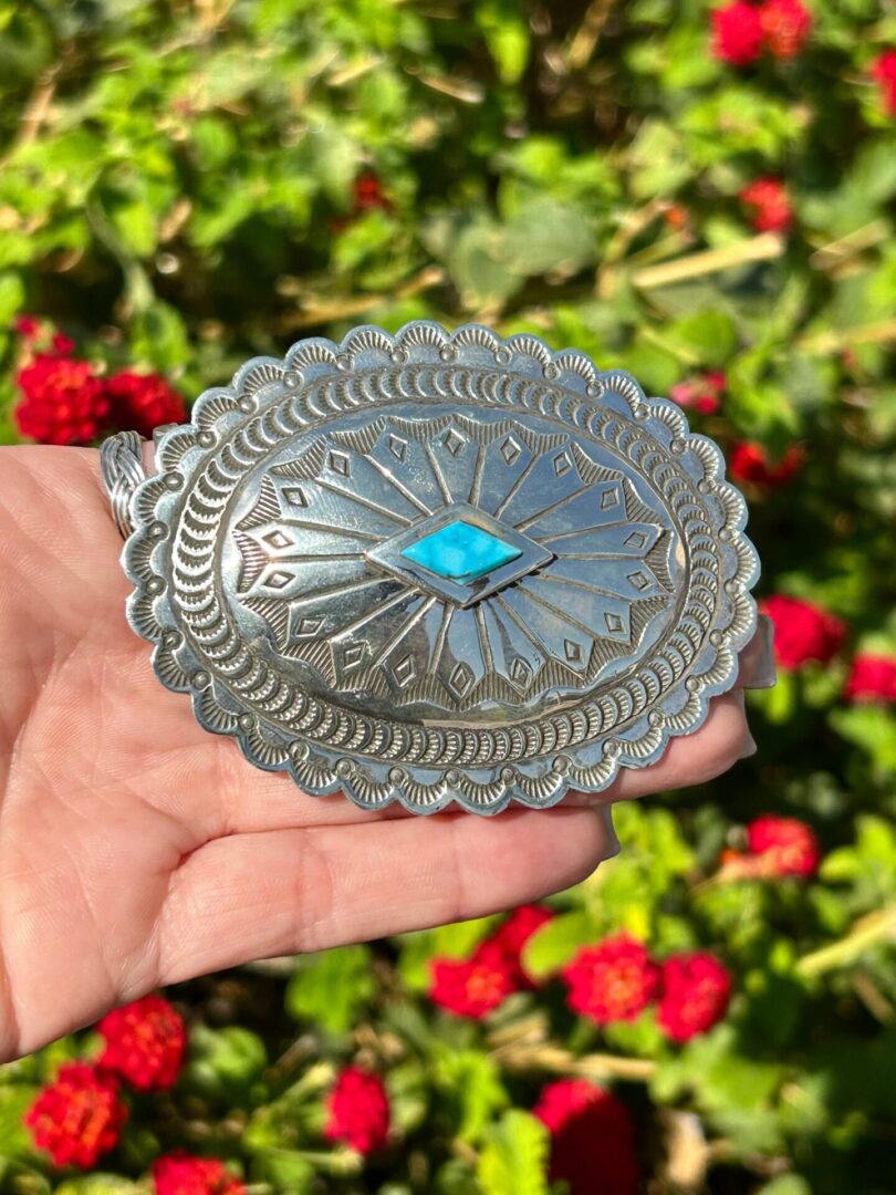 A hand holding a silver belt buckle with a turquoise stone.