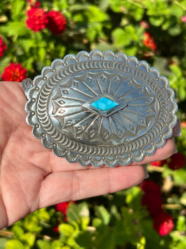 A hand holding a silver belt buckle with turquoise.