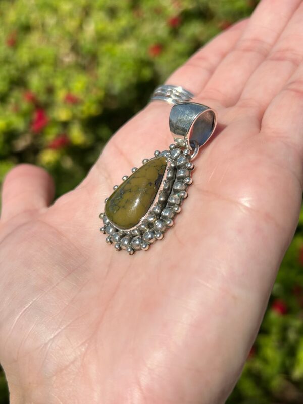 A hand holding a silver pendant with a green stone.