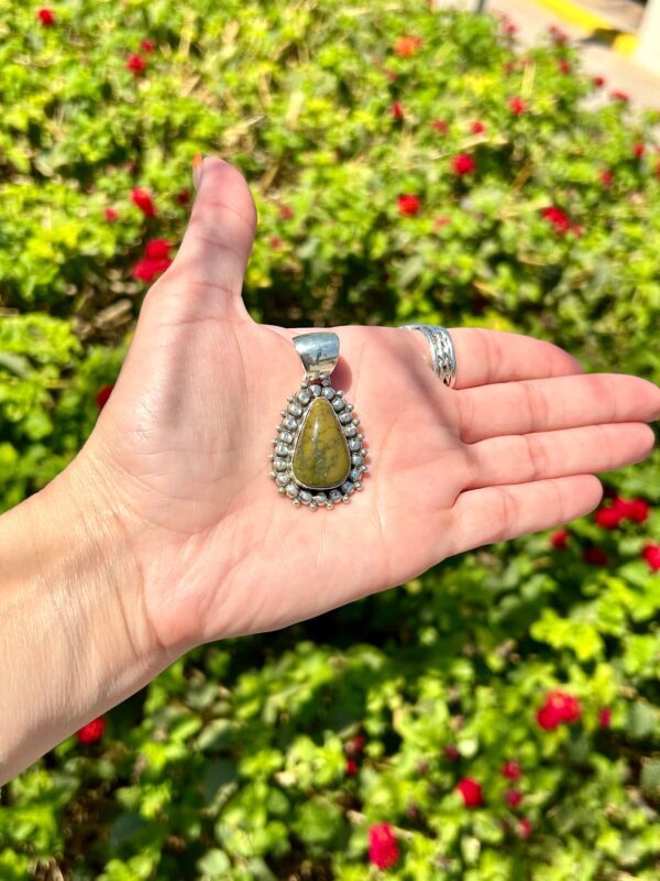 A hand holding a silver pendant with a green stone.