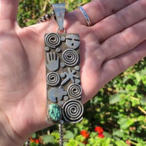 A hand holding a silver pendant with a turquoise stone.