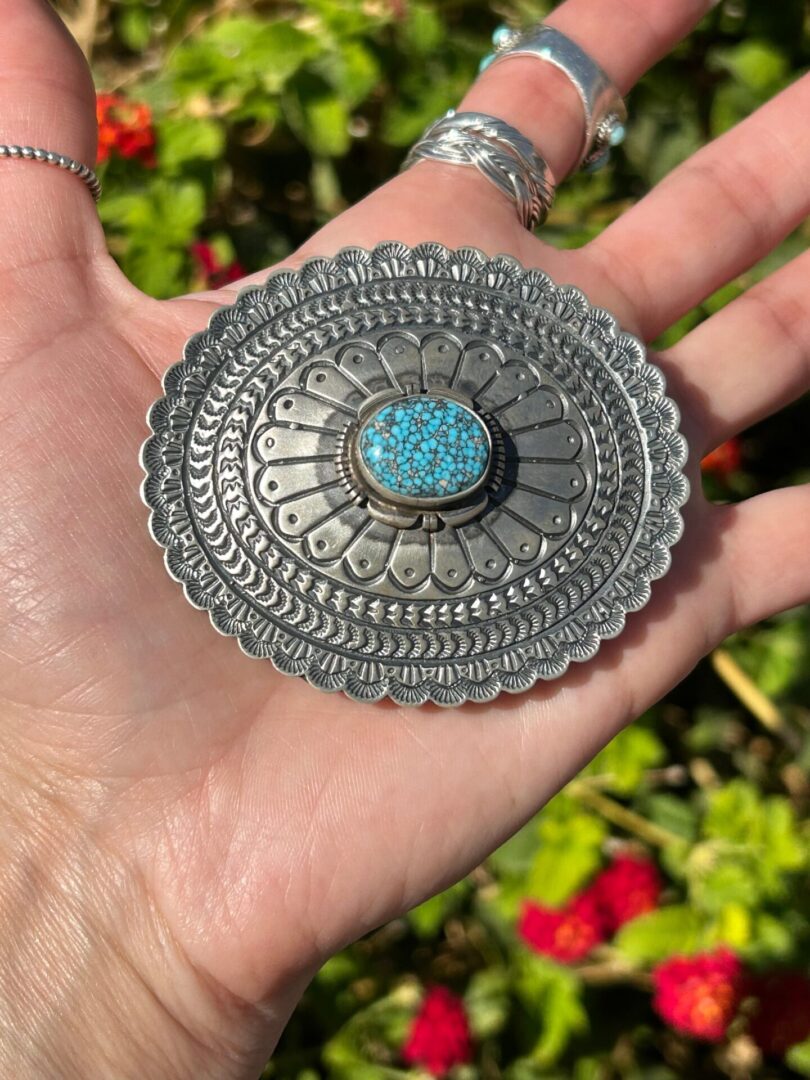 A hand holding a silver buckle with a turquoise stone.