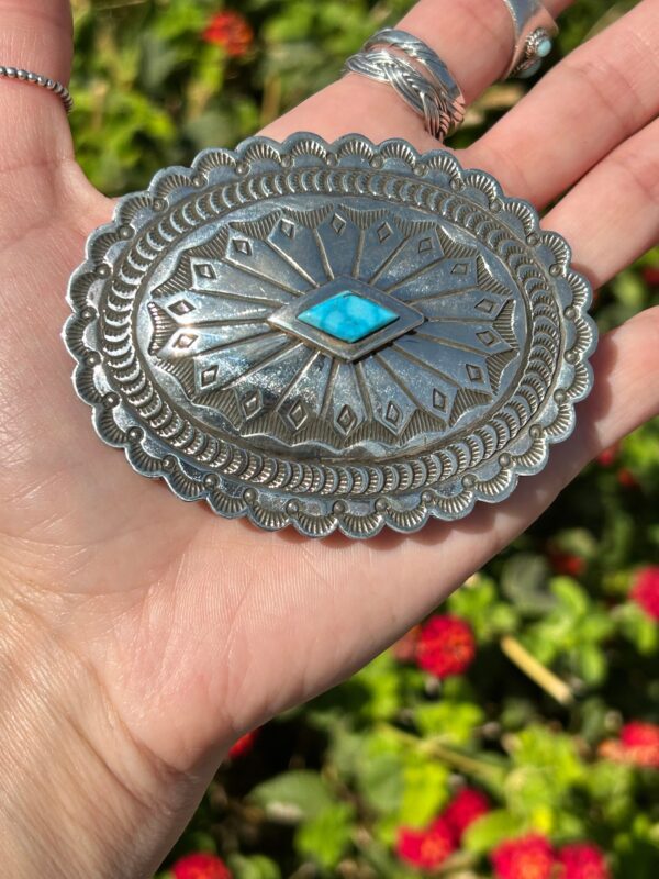 A hand holding a silver buckle with a turquoise stone.