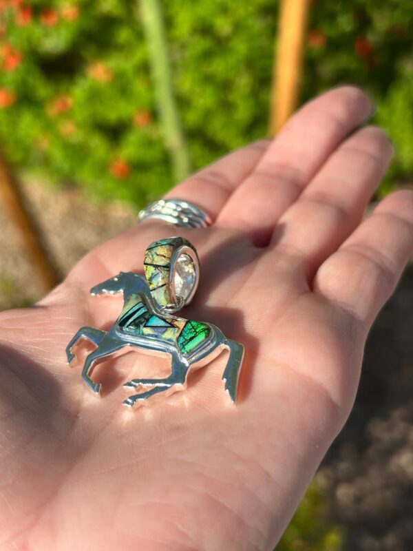 A hand holding a silver horse pendant with green stones.