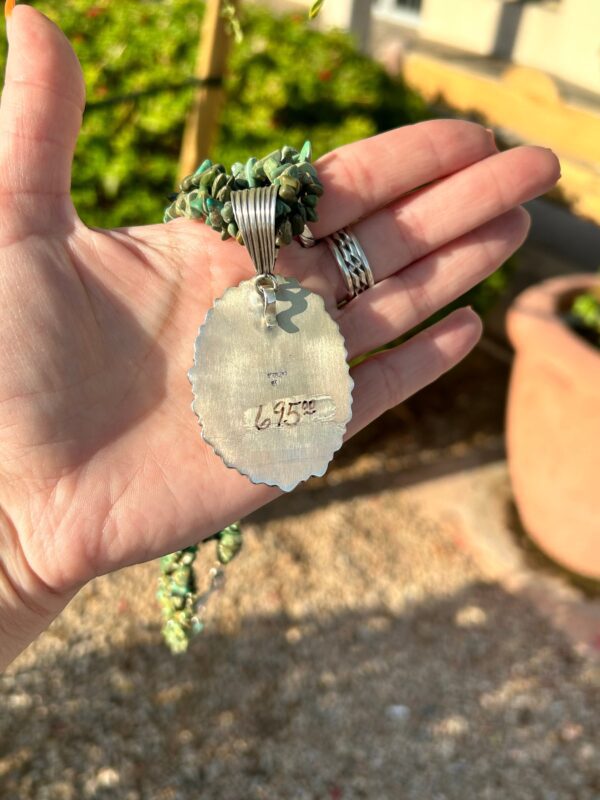 A hand holding a silver pendant with green stones.