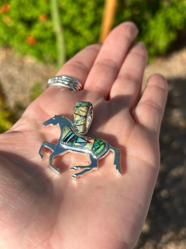 A hand holding a silver horse pendant with colorful inlays.