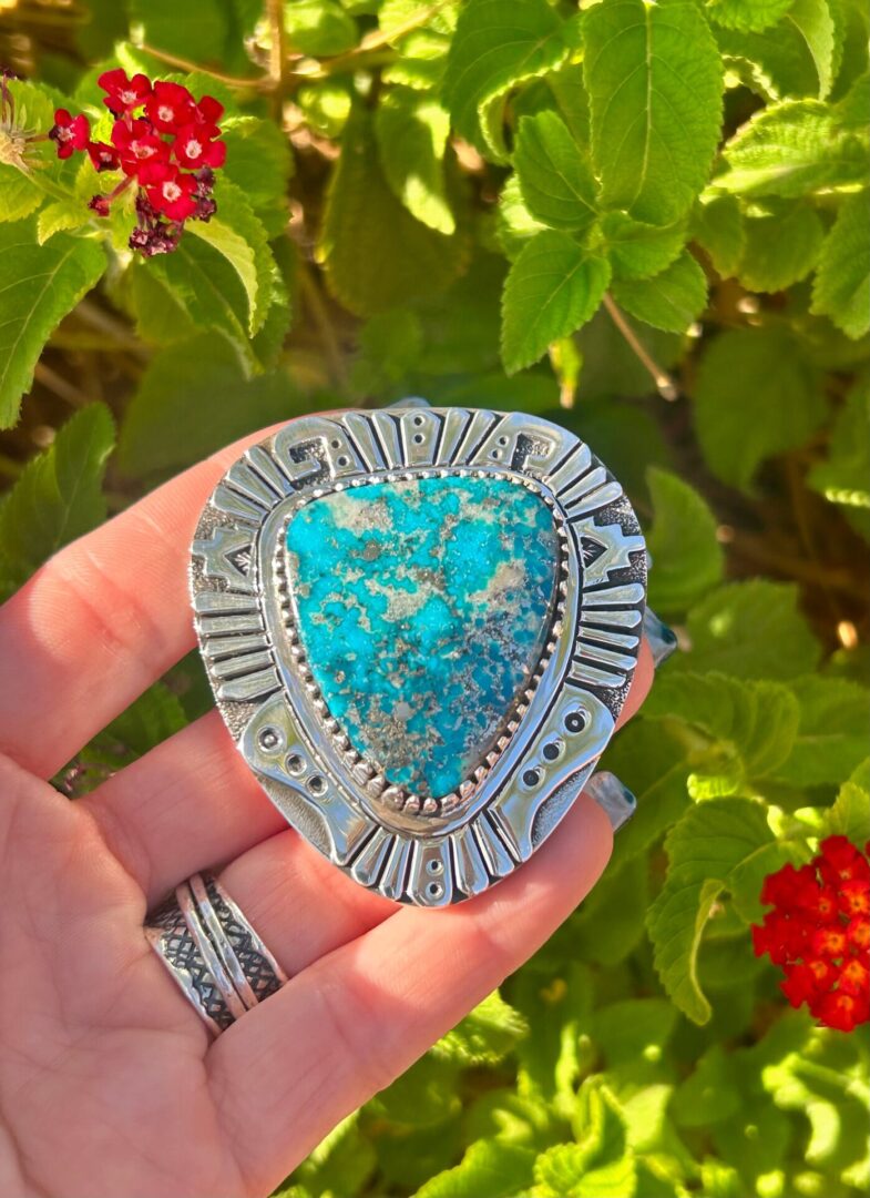 A person holding a turquoise stone brooch in front of flowers.