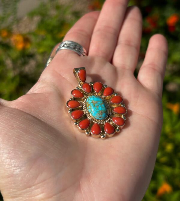 A hand holding a gold pendant with turquoise and coral stones.