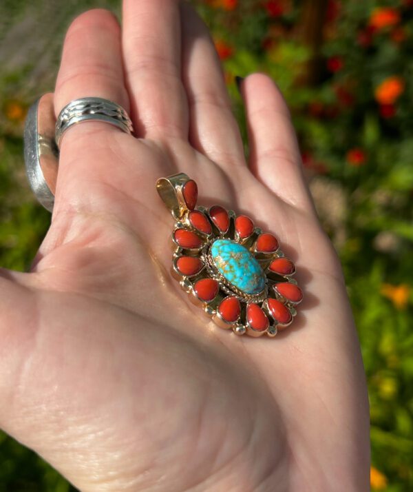 A hand holding a turquoise and coral pendant.