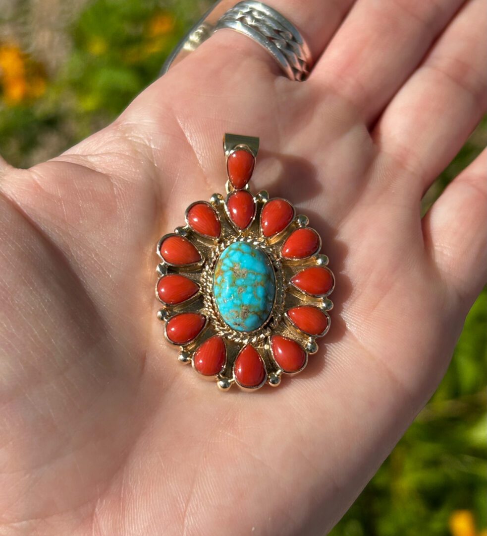 A hand holding a turquoise and coral pendant.