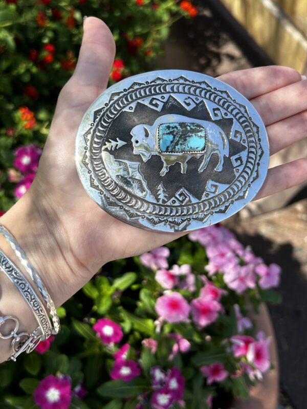 A hand holding a silver belt buckle with a turquoise stone.