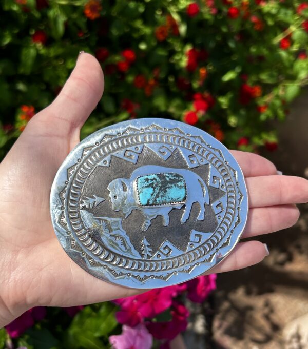 A hand holding a silver and turquoise belt buckle.