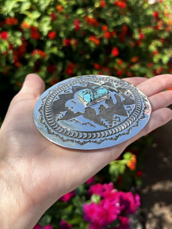 A hand holding a silver and turquoise belt buckle.