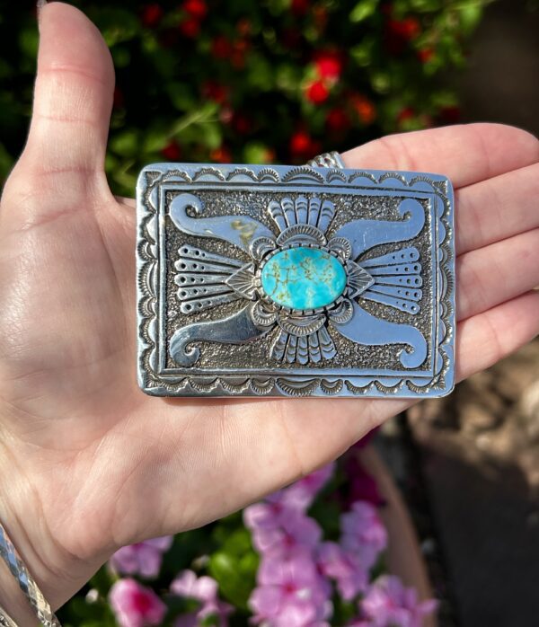 A hand holding a silver belt buckle with a turquoise stone.