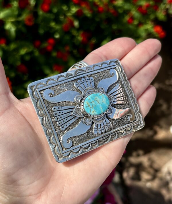 A hand holding a silver belt buckle with a turquoise stone.