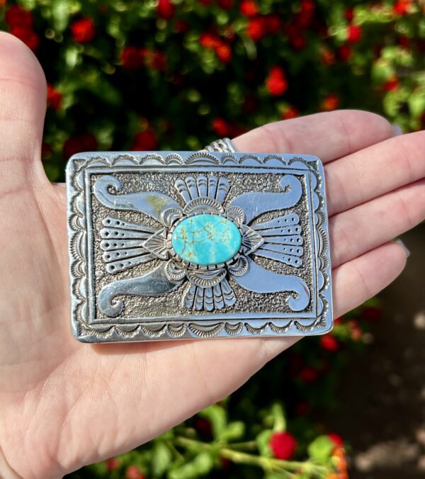 A hand holding a silver and turquoise pendant.