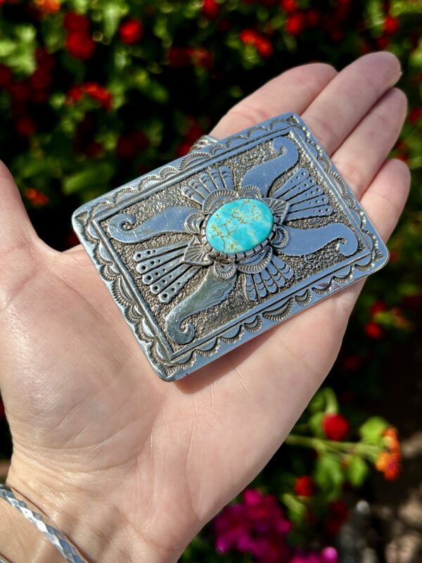 A hand holding a silver belt buckle with a turquoise stone.