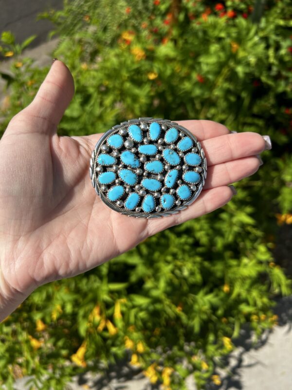 A hand holding a silver and turquoise Native American concha belt buckle.