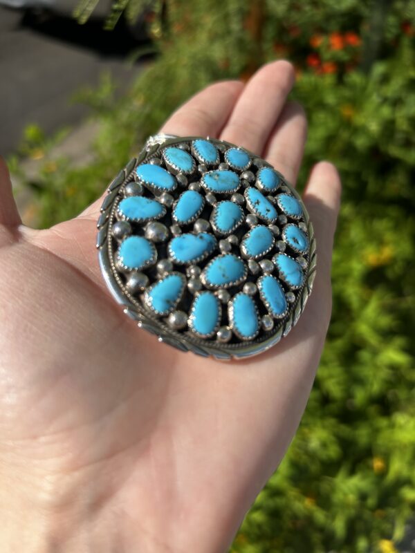 A hand holding a silver and turquoise pendant.