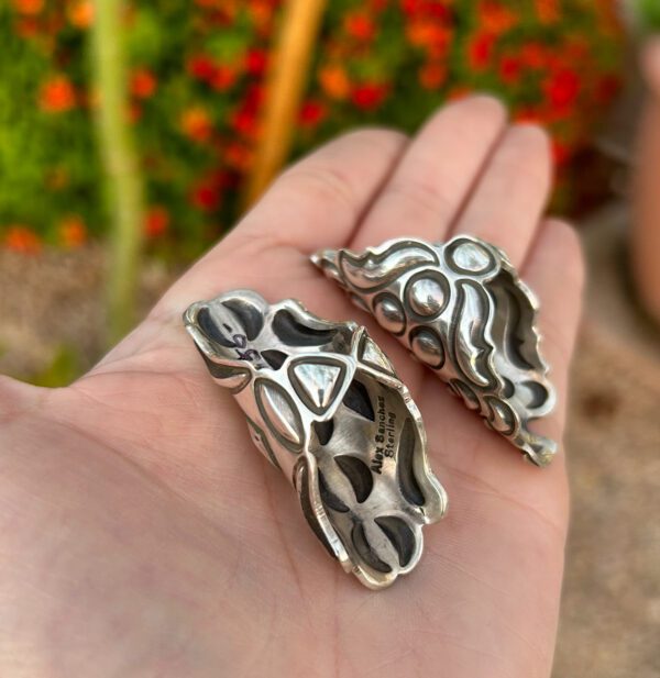 A hand holding two silver concha pendants.