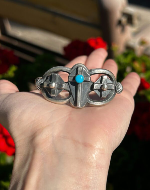 A hand holding a silver and turquoise cross bracelet.